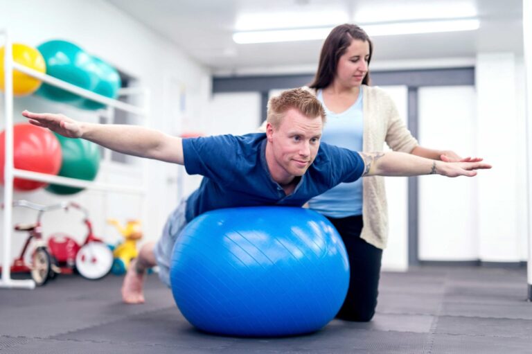 A male physical therapy student in a blue shirt lies face down on a blue exercise ball, extending his arms out to the sides as if flying. A woman stands behind him, providing support and guidance. The room has colorful exercise balls and gym equipment in the background.