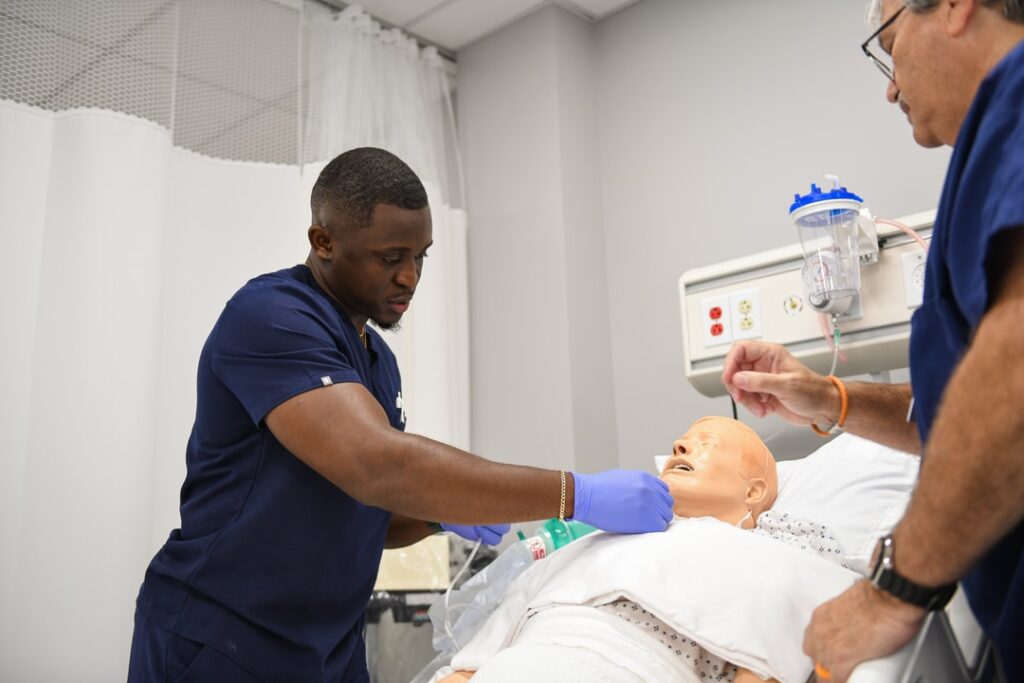 Male Nursing student with instructor checking vital signs on a simulated patient in mock clinical setting.