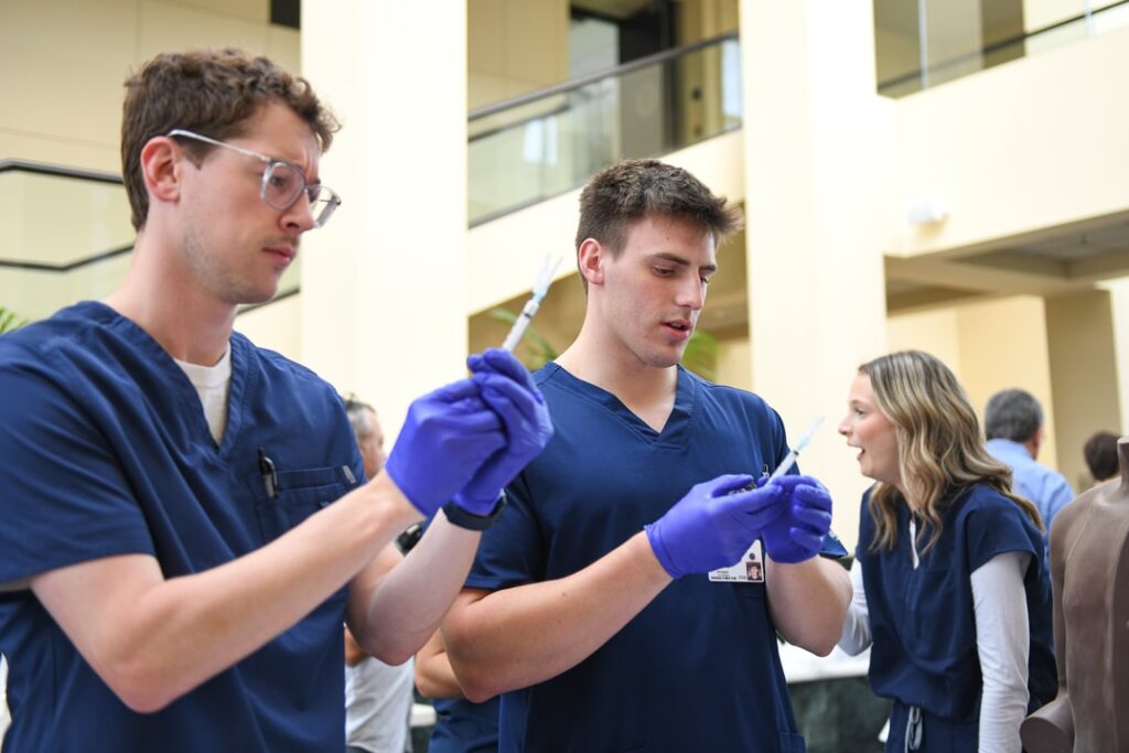 Two male Nursing students in scrubs at skills fair testing syringes for injection