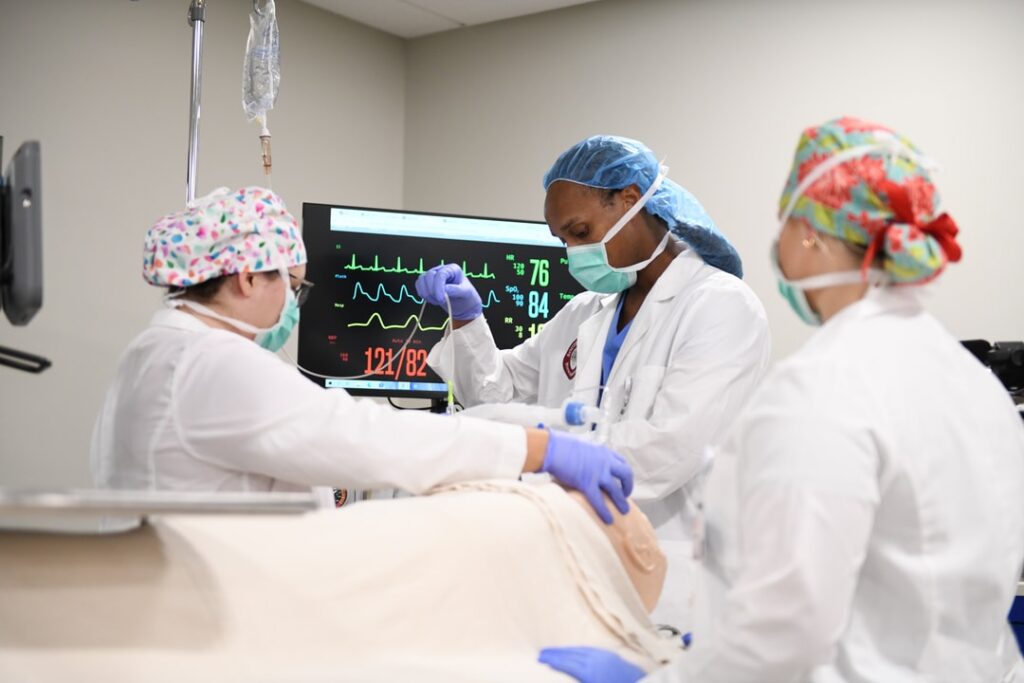 Three Nurse Anesthesia students practicing intubation and anesthesia modeling on simulated patient with vital signs on screen